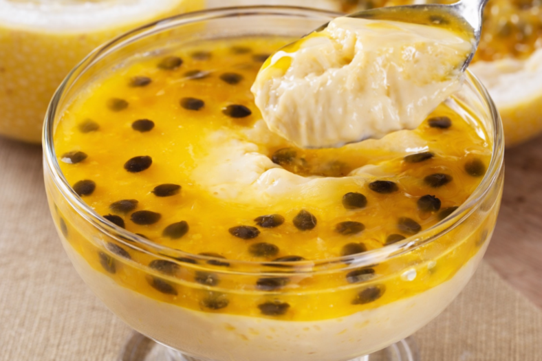 Taça com mousse de maracujá cremosa, coberta com calda sedosa de maracujá e sementes da fruta decorando o topo, em ambiente claro e sofisticado.
