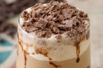 Copo alto com café gelado cremoso, decorado com calda de chocolate nas laterais, chantilly no topo e achocolatado polvilhado, acompanhado de canudo.