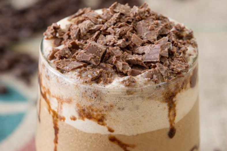 Copo alto com café gelado cremoso, decorado com calda de chocolate nas laterais, chantilly no topo e achocolatado polvilhado, acompanhado de canudo.
