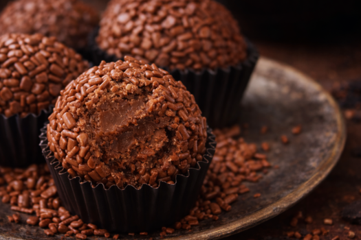 Brigadeiros de café em forminhas marrons, cobertos com chocolate granulado e cacau em pó, dispostos em uma bandeja rústica com grãos de café ao fundo.