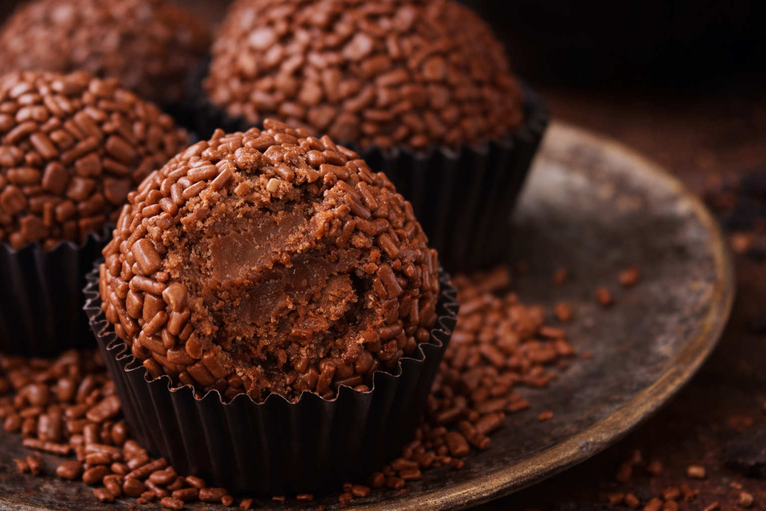 Brigadeiros de café em forminhas marrons, cobertos com chocolate granulado e cacau em pó, dispostos em uma bandeja rústica com grãos de café ao fundo.