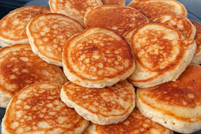 Tareco Bolinhos de frigideira dourados e fofinhos empilhados em um prato, acompanhados de uma xícara de café e um pano de prato xadrez ao fundo.