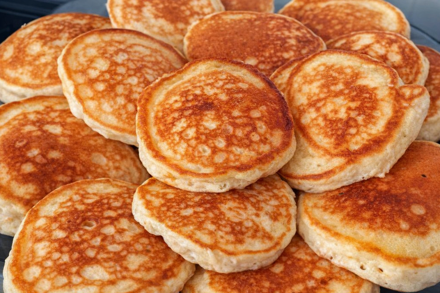 Tareco Bolinhos de frigideira dourados e fofinhos empilhados em um prato, acompanhados de uma xícara de café e um pano de prato xadrez ao fundo.