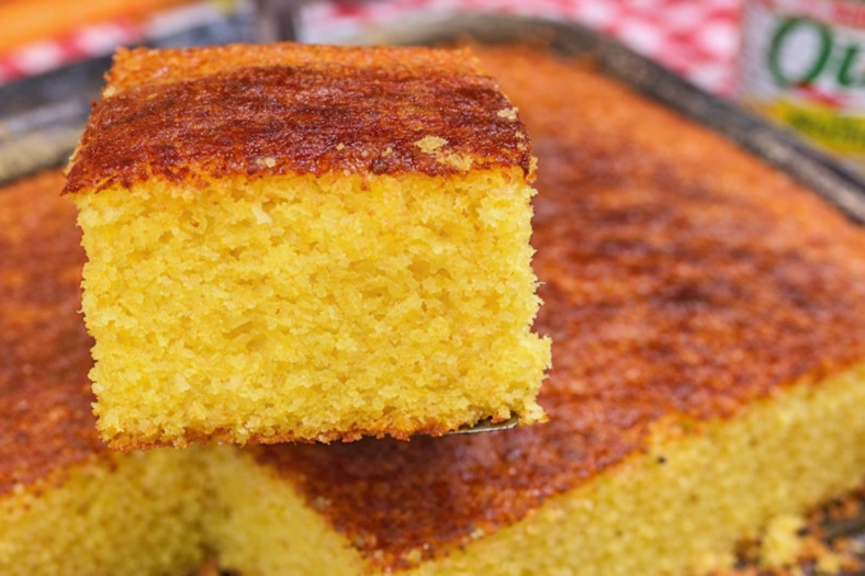 Bolo de milho cremoso em forma de buraco no meio, com superfície dourada e fatia servida em prato branco ao lado de uma xícara de café.