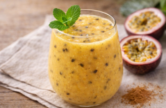 Copo de batida de maracujá cremosa com sementes e folhas de hortelã, sobre superfície de madeira com maracujá cortado ao lado.