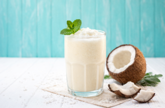 Copo de batida de coco cremosa com pedaços de coco ao redor, sobre superfície branca com fundo azul turquesa.
