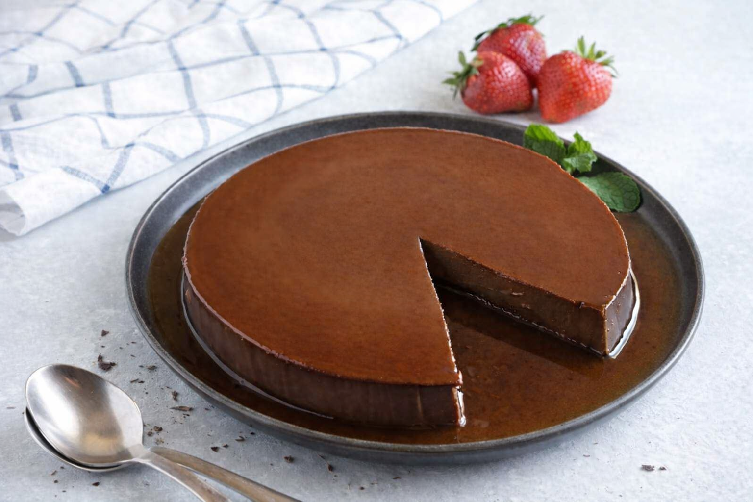 Flan de chocolate brilhante em prato escuro com morangos frescos e folhas de hortelã ao lado.