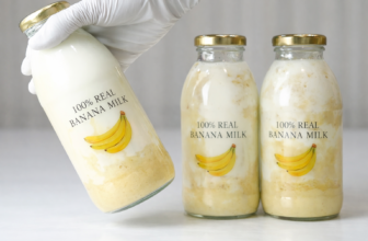 Três garrafas de vidro de leite de banana com tampas douradas e rótulo “100% REAL BANANA MILK” sobre superfície branca.